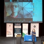 Duża scena Kieleckiego Centrum Kultury. Na scenie przemawia prowadząca konferencję. W tle sceny na planszy projekcyjnej wyświetlony jest online Pan Paweł Słowik przyjmujący gratulacje od prowadzącej konferencję.