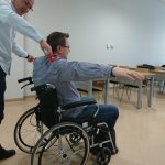 Na zdjęciu w sali wykładowej dwóch mężczyzn jeden na wózku inwalidzkim z rozłożonymi rękoma na boki, drugi mężczyzna z miarką mierzący rozstaw jego rąk. Mężczyźni uczestniczą w szkoleniu pt. „Dostępność przestrzeni akademickiej dla osób z niepełnosprawnościami”.