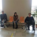 Zdjęcie korytarza na którym przebywają trzy osoby. Pierwsza osoba po lewej to mężczyzna siedzący na wózku inwalidzkim obok niego kobieta na krześle i kolejno kobieta na wózku inwalidzkim. Osoby są uczestnikami szkolenia, doświadczają niepełnosprawności- rozmawiają ze sobą.