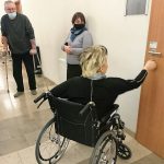 Zdjęcie korytarza na którym przebywają trzy osoby. Pierwsza osoba z prawej to kobieta siedząca na wózku inwalidzkim, próbująca otworzyć drzwi do sali, po jej lewej stronie przyglądająca się kobieta oraz mężczyzna o kulach. Wszystkie osoby biorą udział w szkoleniu.