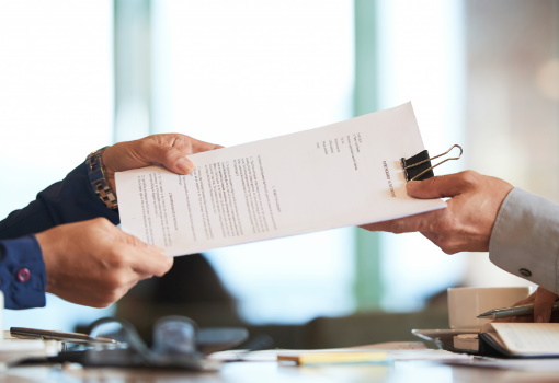 W pomieszczeniu biurowym mężczyzna z prawej strony w szarej koszuli przekazuje mężczyźnie po lewej stronie w granatowej koszuli plik dokumentów przytrzymanych spinaczem klipsowym.