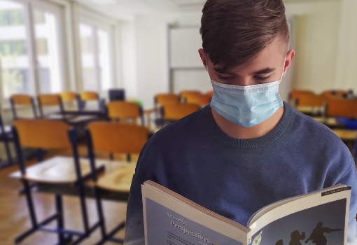 Student stoi w klasie tyłem do tablicy. Ma założoną na nos i usta maseczkę i czyta otwartą książkę. Za nim widać krzesełka, okna i tablicę.