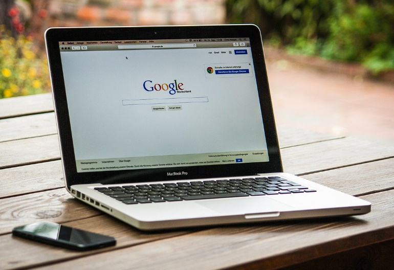 Otwarty laptop firmy Apple lezy na drewnianym stole. Obok niego leży telefon komórkowy.