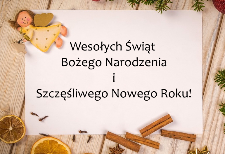 Na drewnianym blacie leży biała kartka z życzeniami świątecznymi. Wokół niej są ozdoby i elementy związane z Bożym Narodzeniem: figurka aniołka, suszone plastry pomarańczy, laski cynamonu, goździki i zielone gałązki.