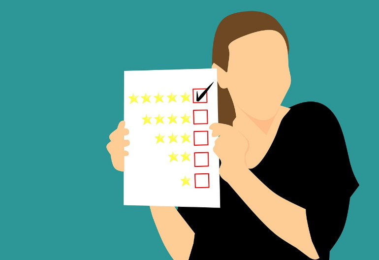 Kobieta ubrana w ciemną bluzkę trzyma kartkę prezentując jej treść. Na kartce są oceny od 1 do 5 gwiazdek i zaznaczony głos przy 5 gwiazdkach.
