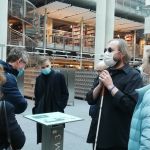 Hol Biblioteki Uniwersytetu Warszawskiego, w którym uczestnicy wizyty słuchają przewodnika. Ma on na sobie ciemne okulary i trzyma oburącz białą laskę.