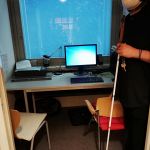 Przewodnik w ciemnych okularach i z białą laską stoi w boksie, w którym jest biurko z komputerem, skanerem i akcesoriami.