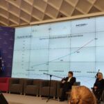 Prelegentki omawiają prezentację dotyczącą realizacji projektu. W tle duży slajd z podaną liczbą przeszkolonych pracowników. Dwoje moderatorów przysłuchuje się wystąpieniu, jedna osoba tłumaczy na Polski Język Migowy.