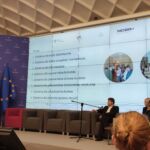 Prelegentki omawiają prezentację dotyczącą realizacji projektu. W tle duży slajd z wymienionymi grupami szkoleniowymi. Dwoje moderatorów przysłuchuje się wystąpieniu, jedna osoba tłumaczy na Polski Język Migowy.