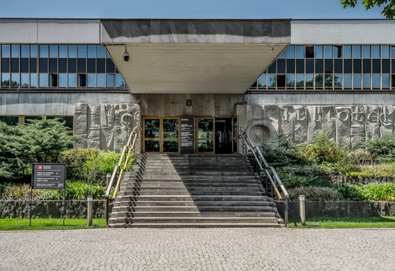 Wejście do głównego gmachu Biblioteki Narodowej, do którego prowadzą schody. Przed budynkiem rośnie trawa oraz krzewy. Schody są zadaszone.