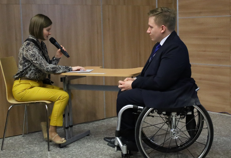 Michał Wrona siedzi na wózku inwalidzkim i słucha wypowiedzi Karoliny Malickiej czytającej notatki brailem.