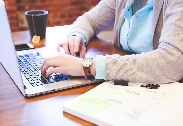 Na stole rozłożony laptop, z którego korzysta kobieta ubrana w koszulkę i bluzkę. Obok stoi kubek i leży telefon i zeszyt.