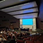 Siedzący uczestnicy konferencji w auli słuchają wystąpienia prelegentki, za nią znajduje się duży ekran.