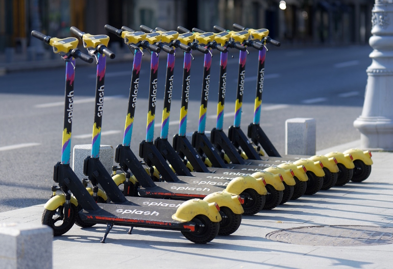 Przy krawędzi miejskiej ulicy w równych odstępach stoi dziesięć hulajnóg elektrycznych.
