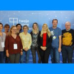 Pracownicy Centrum Wsparcia pozują do zdjęcia w gronie przedstawicieli innych uczelni. W tle plansza organizatora z logo.