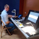 Mgr Sebastian Grabek siedzący przy dużym stole prezentuje działanie urządzeń wspomagających pracę osób z niepełnosprawnościami.