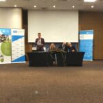 Sala konferencyjna. Za dwoma długimi stołami siedzą uczestnicy Zjazdu. Przy trzecim siedzą trzy osoby, czwarta stoi z mikrofonem w ręku i przemawia.