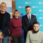 Troje pracowników Centrum Wsparcia i współorganizator Konferencji pozują do zdjęcia w sali konferencyjnej. Za nimi stoją roll-upy.