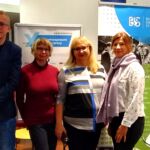 Dwoje pracowników Centrum Wsparcia i dwie przedstawicielki innych uczelni uśmiechają się i pozują do zdjęcia w pomieszczeniu. Za nimi stoją roll-upy.