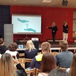 Warsztaty z języka migowego. W tyle sali na podwyższeniu trzy panie prowadzące warsztaty, opowiadają i migają. Z przodu widownia.